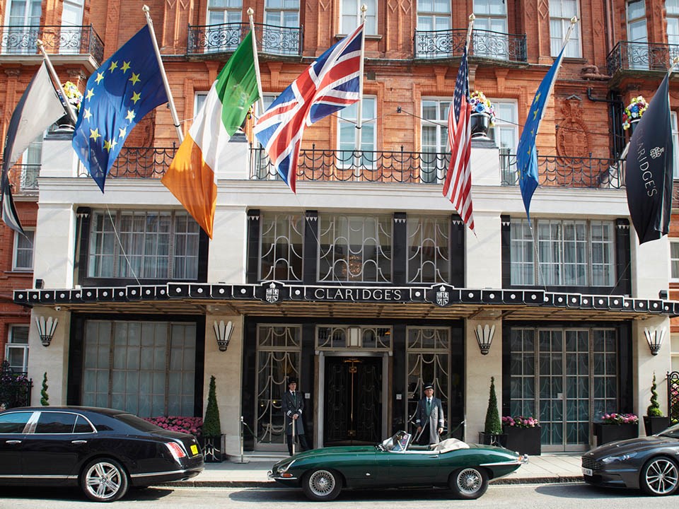 A view of the entrance outside Claridge's Hotel, with doorkeeper, flags and parked cars outside.