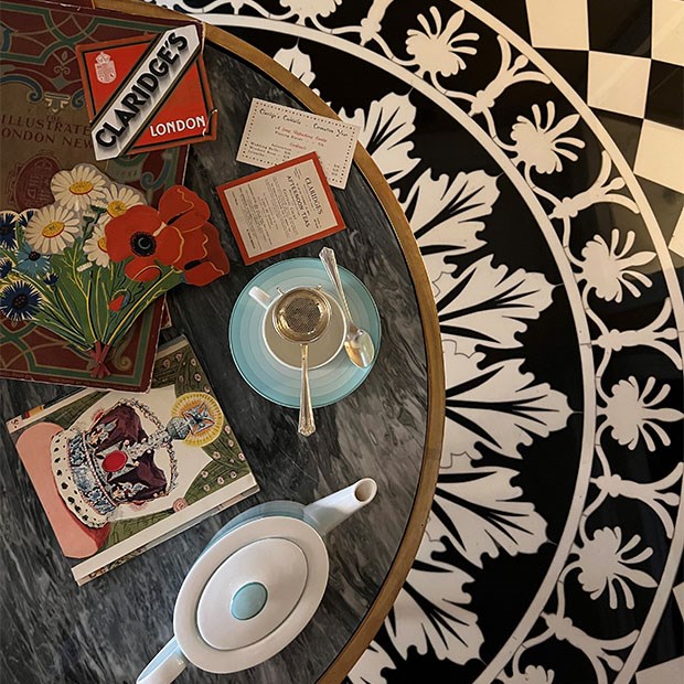 Marble table with Claridge’s memorabilia, vintage menus, and postcards alongside a teapot, cup, and saucer set on a patterned floor.