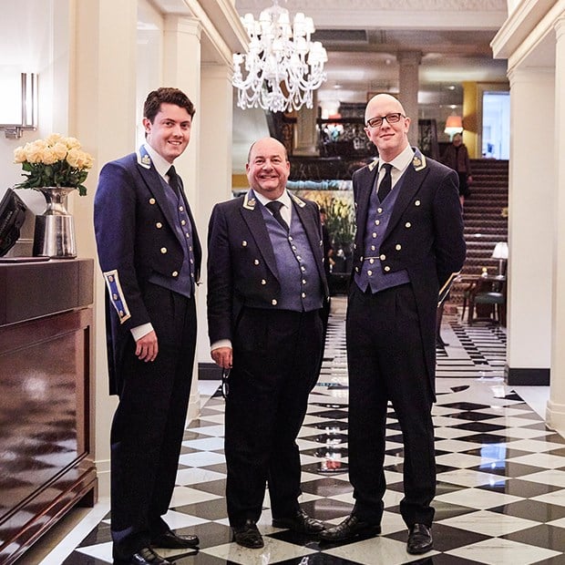 Three smiling concierges stand and pose in the corridor of Claridge's Hotel.