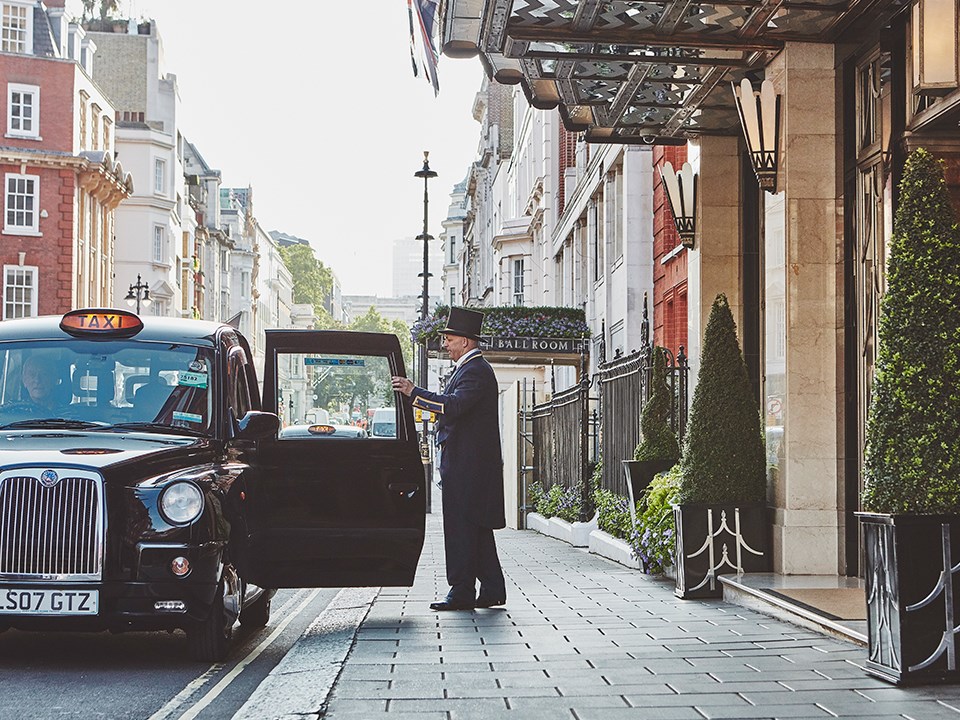 A smiling doorman opening the door of a taxi to welcome guests, outside the Claridge's Hotel.