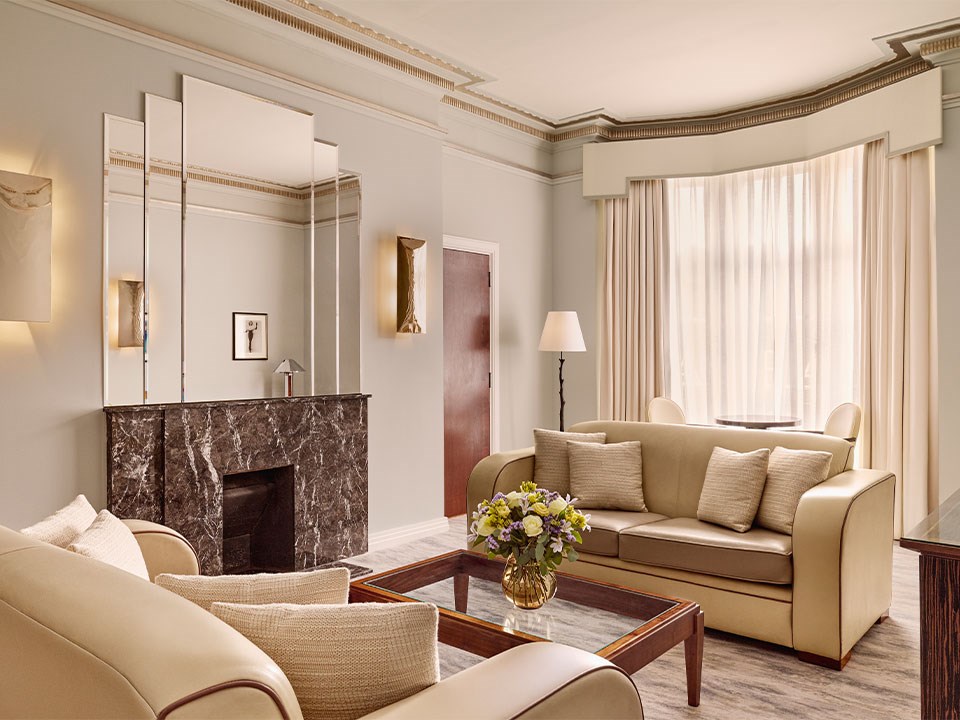 Elegant hotel suite living area with cream leather sofas, a dark marble fireplace, mirrored wall panels, and a large bay window dressed with sheer curtains.