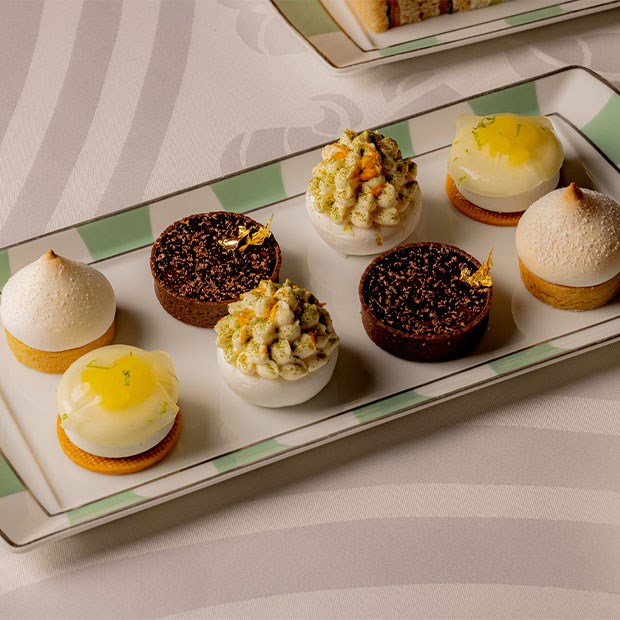 A meticulously arranged selection of delicate pastries, featuring lemon tarts, chocolate ganache bites with gold leaf, and pistachio-adorned meringues, elegantly presented on a striped Claridge’s plate.