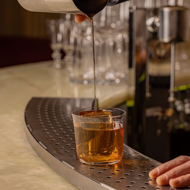 A golden-hued drink being gracefully poured over a single clear ice cube in a modern glass, capturing a moment of refinement.