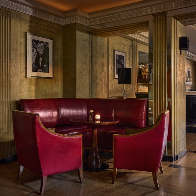 Intimate corner seating at Claridge’s Bar with red leather banquette, two matching armchairs, and a small round table, set against gold-toned walls with framed black-and-white photography.
