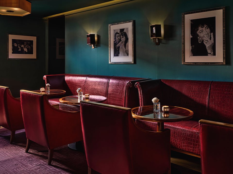 An intimate lounge area with deep red leather seating, dark green walls, low-lit sconces, and black-and-white celebrity portraits, creating a moody, sophisticated atmosphere.