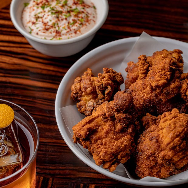A bowl of crispy, golden-brown fried chicken pieces served alongside a creamy dipping sauce sprinkled with herbs, paired with a cocktail garnished with a citrus twist on a dark wooden table.