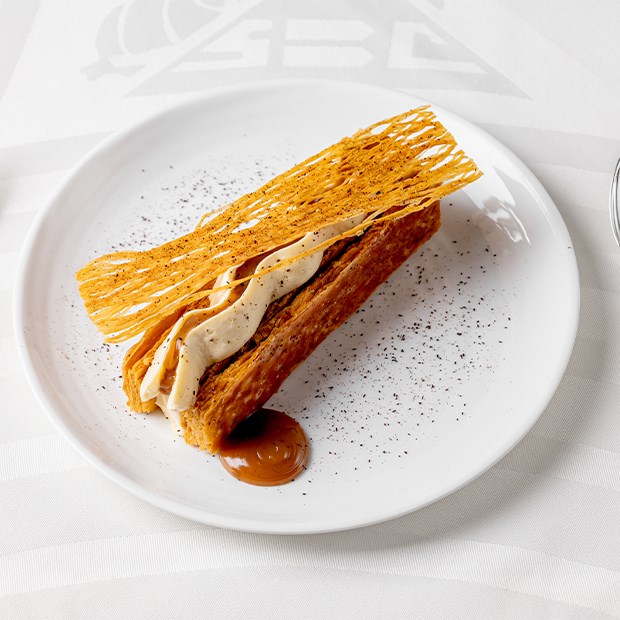 A beautifully plated dessert featuring layers of delicate, crisp pastry, a rich cream filling, and a thin, lacy garnish, artfully drizzled with a caramel sauce on a pristine white plate.