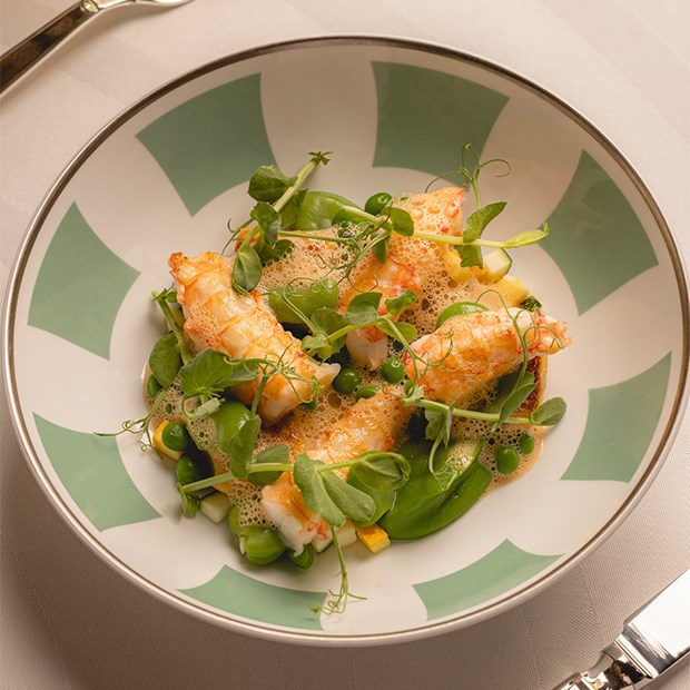 A gourmet dish featuring tender pieces of lobster or langoustine, garnished with fresh green peas, microgreens, and a light foam sauce, elegantly plated on a patterned dish.