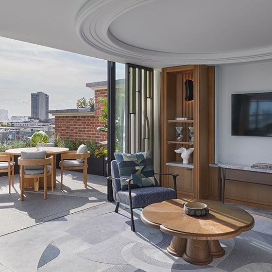 Modern suite living area with curved ceiling detail, a round wooden coffee table, and a blue armchair. Glass doors open onto a private terrace with dining furniture and city skyline views. A built-in wooden display cabinet and wall-mounted TV complete the interior décor.