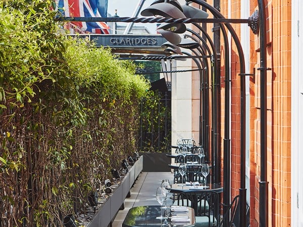 Chic outdoor dining terrace at Claridge’s, featuring black metal tables set with wine glasses and cutlery. The terrace is bordered by tall green hedges on one side and a red-brick building with curved black awnings on the other, with Union Jack flags visible in the background.