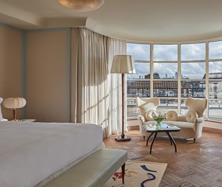 Elegant hotel suite with parquet flooring, a large bed in the foreground, and a curved wall of floor-to-ceiling windows revealing a city skyline. The sitting area features two sculptural white armchairs, a glass coffee table with fresh flowers, and a tall floor lamp beside sheer curtains.