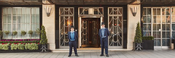 Two doormen in navy uniforms and top hats stand smiling at the entrance of Claridge’s, framed by Art Deco-style doors and potted greenery.