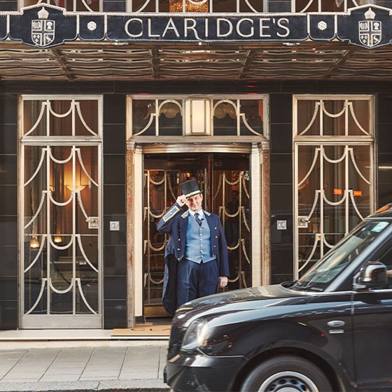 A doorman in a navy uniform and top hat tips his hat outside the Claridge’s entrance as a black taxi passes by.