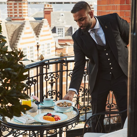 A hotel staff member in a black suit sets a bowl of granola on a balcony table with fruit and tea, overlooking red-brick and white-trimmed rooftops.