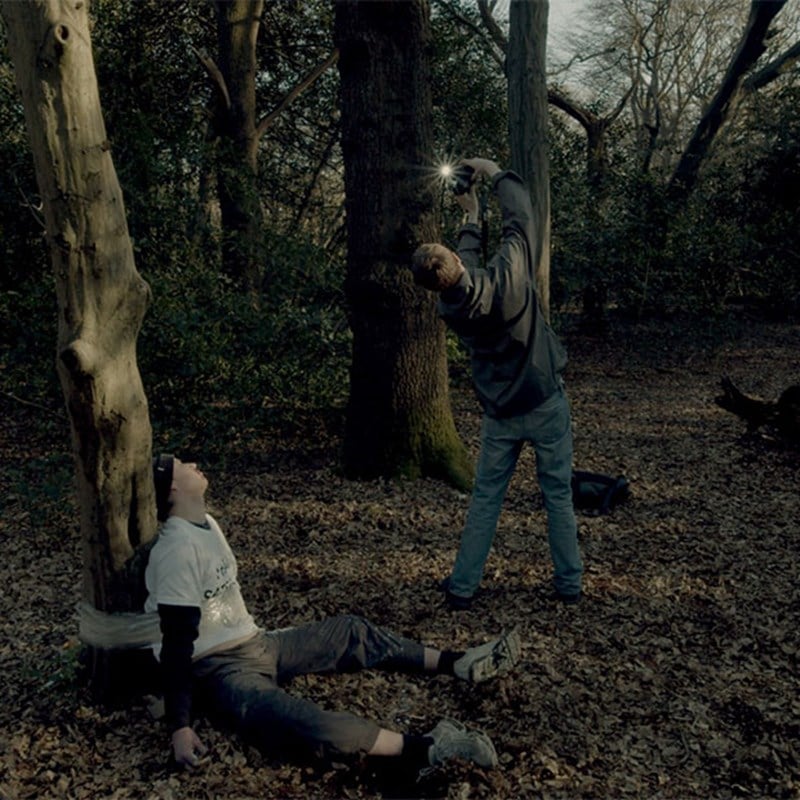Man tied to a tree in the woods with another man standing nearby, raising a camera flash.