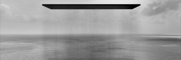 A black-and-white seascape showing a vast, calm ocean beneath a cloudy sky. A large, flat black rectangle hovers silently above the water, its shadow faintly reflected below, creating an otherworldly, abstract atmosphere.