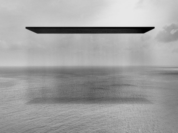 A black-and-white seascape showing a vast, calm ocean beneath a cloudy sky. A large, flat black rectangle hovers silently above the water, its shadow faintly reflected below, creating an otherworldly, abstract atmosphere.