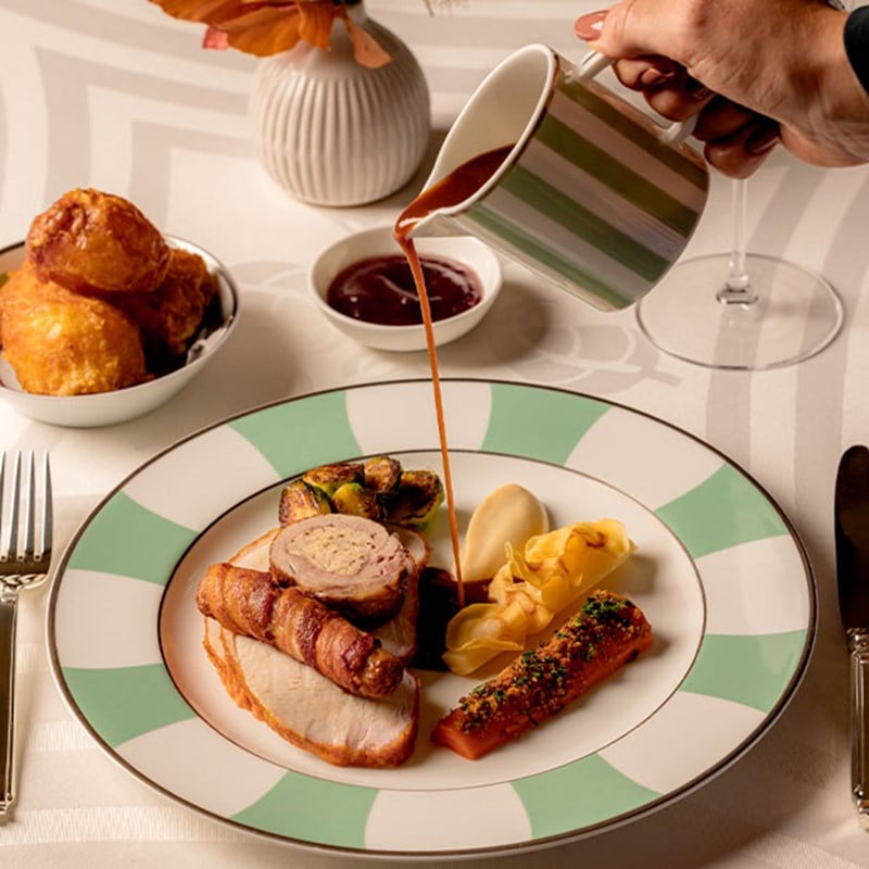 A sophisticated roast dish featuring turkey wrapped in pancetta, a variety of vegetables, and a rich sauce being elegantly poured over the plate, paired with traditional Yorkshire puddings.