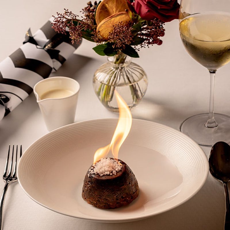 A flaming Christmas pudding elegantly presented in a white bowl, paired with a small jug of cream, a glass of wine, and festive floral decor in the background.