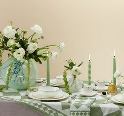 A charming table arrangement featuring a turquoise speckled vase filled with white ranunculus and trailing greenery, accompanied by gold-edged white china, mint-green twisted candles, and a geometric green-and-white tablecloth, radiating refined elegance.
