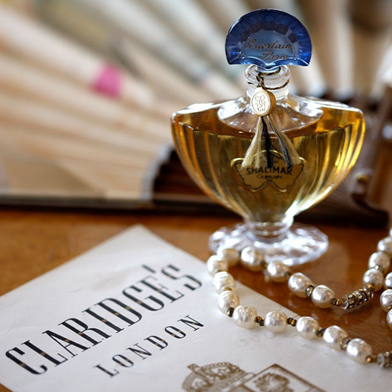 A classic perfume bottle of Shalimar by Guerlain with a pearl necklace and a Claridge's paper on a wooden surface, reflecting an elegant atmosphere.