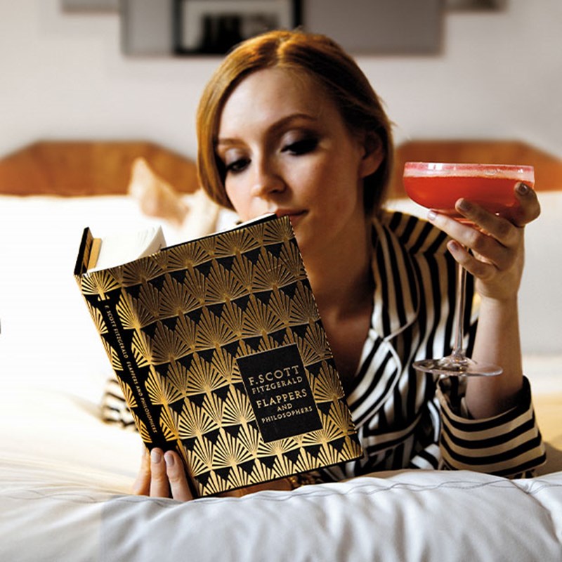 A woman in striped pajamas holding a pink cocktail while reading F. Scott Fitzgerald's