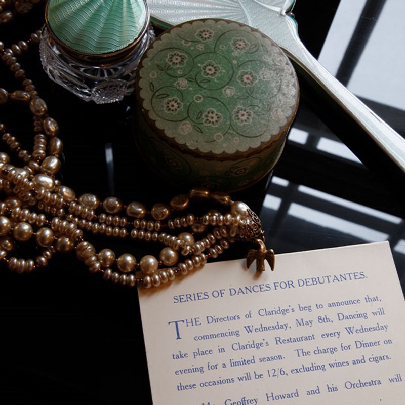 A collection of vintage items such as pearls, a decorative container, and a card announcing a series of dances at Claridge’s, evoking the glamour of the past.