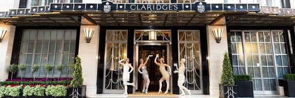 Four dancers in sparkling flapper dresses striking poses outside the iconic entrance of Claridge’s, with British flags flying above.