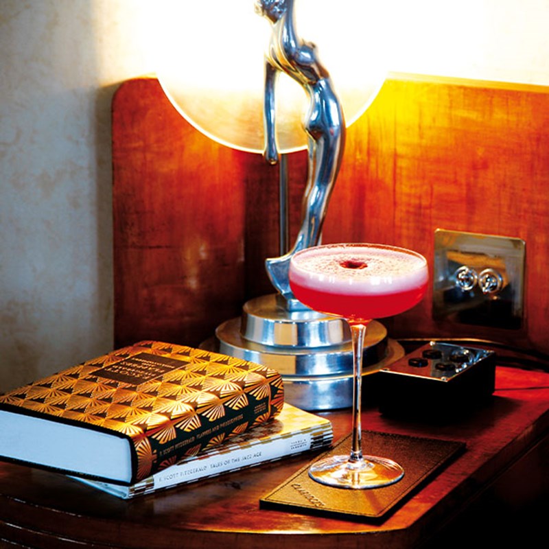 Pink cocktail on Art Deco bedside table with sculptural lamp and patterned book.