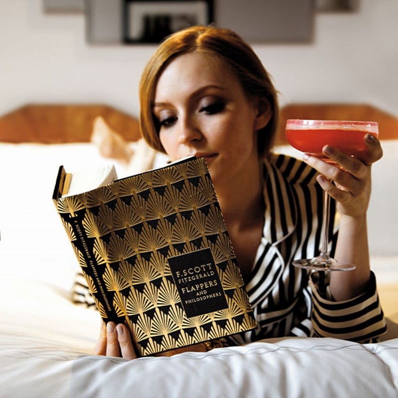 Woman in striped pyjamas reading F. Scott Fitzgerald and holding a cocktail in bed.