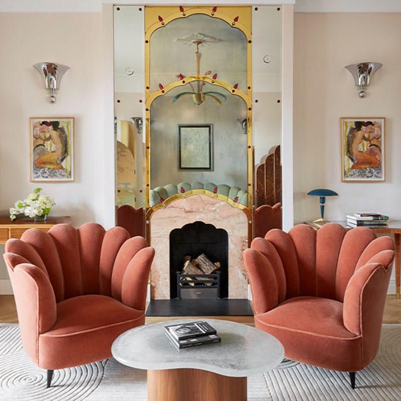 Two scalloped velvet chairs facing fireplace with mirrored Art Deco detailing and pastel art.