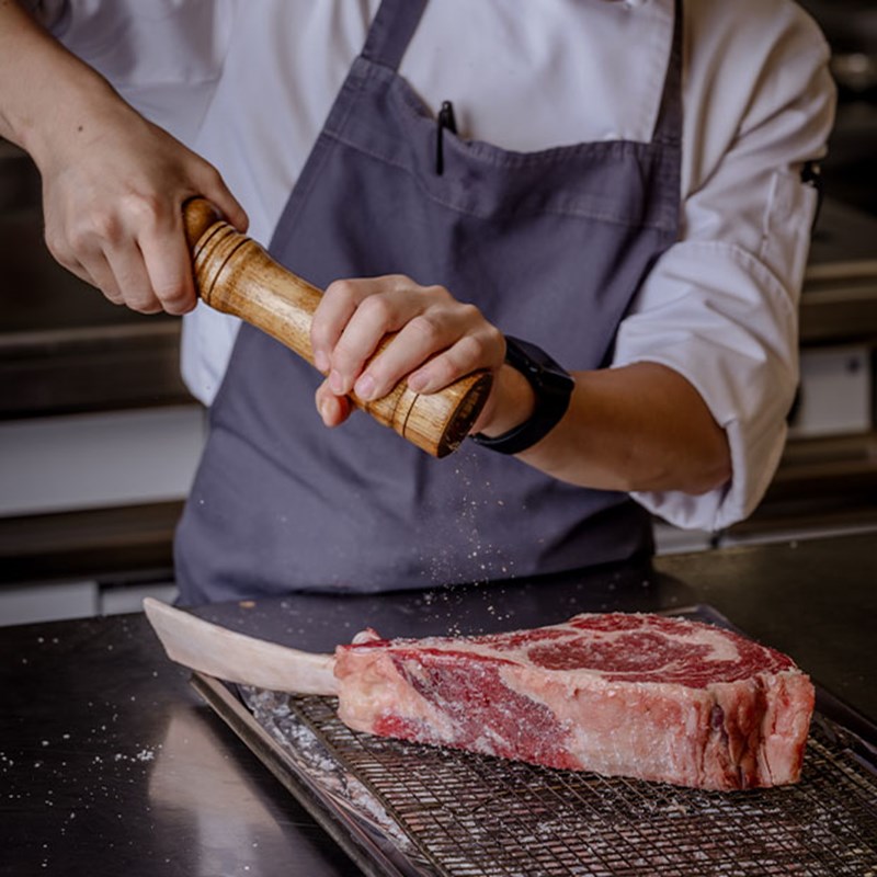 A chef in a crisp white uniform grinds fresh black pepper over a thick tomahawk steak, preparing it for the grill with precision and care.