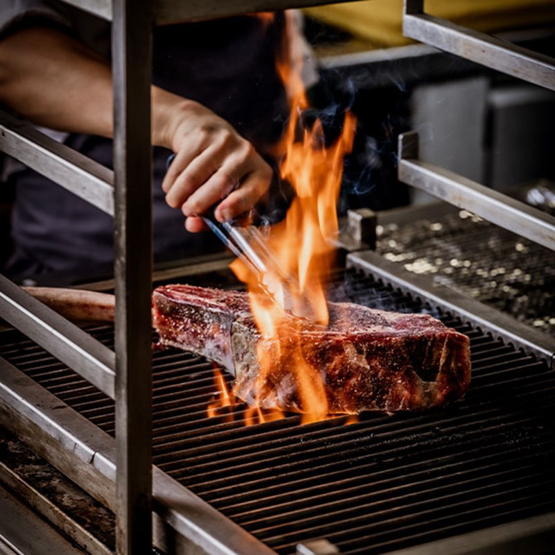 Flames rise as a chef expertly sears a tomahawk steak over an open grill, creating a rich, smoky char that locks in the flavour.