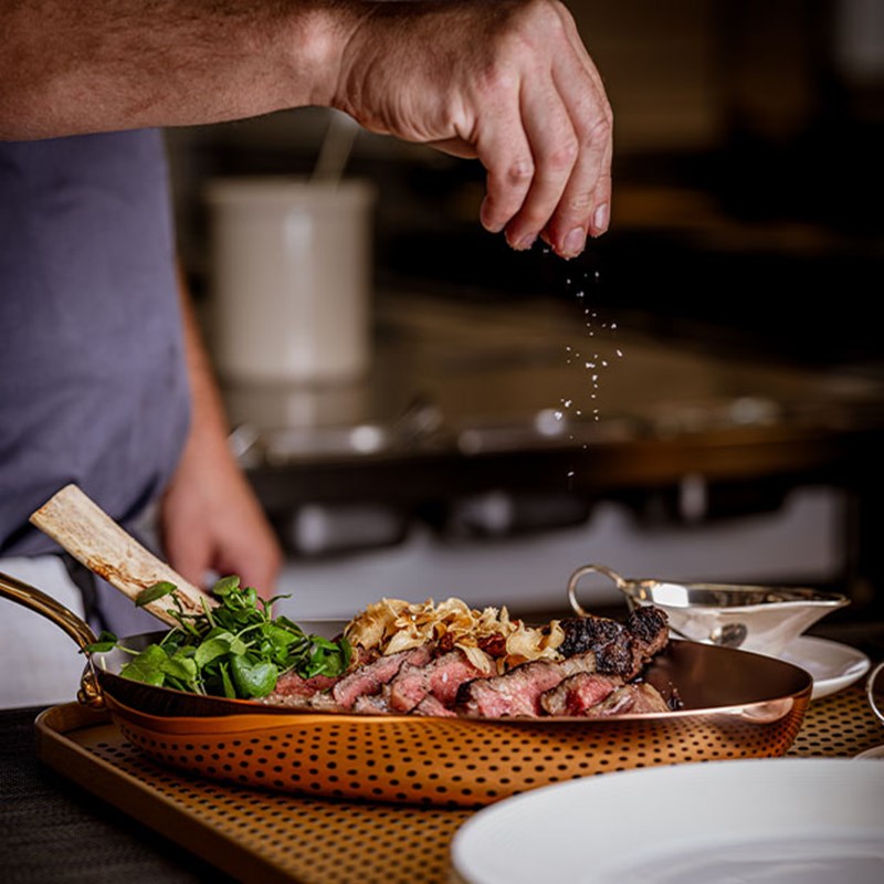 A chef sprinkles sea salt over a perfectly cooked tomahawk steak, served in a gleaming copper pan alongside crispy shallots and fresh watercress, ready to be plated.