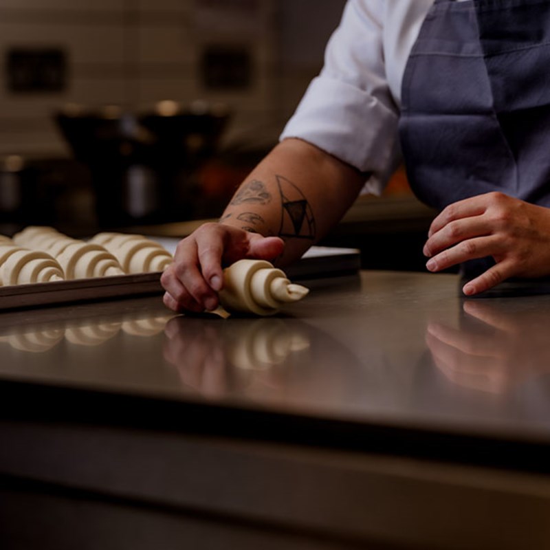 A pastry chef’s hands carefully roll croissant dough on a stainless steel worktop, poised for baking, in a setting where precision meets artistry.