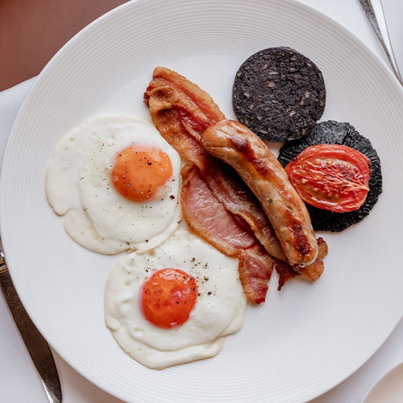 A meticulously plated full English breakfast, featuring two fried eggs with bright yolks, crispy bacon, a plump sausage, grilled tomato, black pudding, and a side of toast.