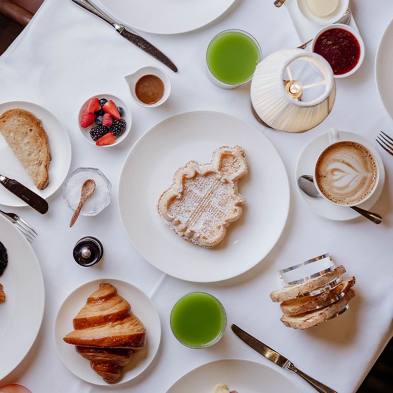 A beautifully arranged breakfast table featuring a Claridge’s crest-shaped waffle dusted with powdered sugar, a golden croissant, fresh fruit, slices of toast, green juice, a cappuccino with latte art, and delicate accompaniments on fine china.