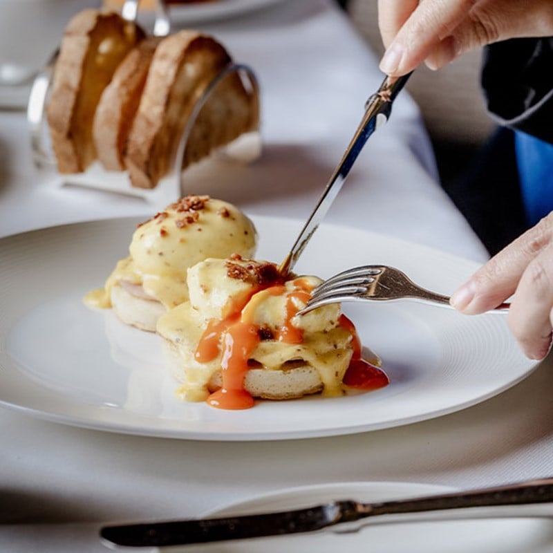 A guest elegantly cuts into a plate of eggs Benedict, the rich hollandaise sauce cascading over poached eggs and English muffins, accompanied by crispy bacon and vibrant tomato sauce.