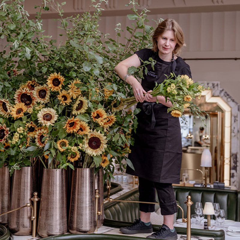 A florist in a black apron carefully arranges fresh sunflowers and lush greenery in tall bronze vases, creating a striking centrepiece for Claridge’s dining room.