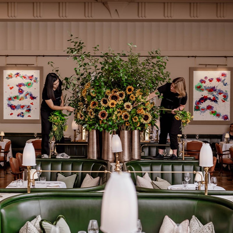 Two florists stand atop green banquettes, meticulously styling an opulent sunflower arrangement in the heart of Claridge’s, with vibrant contemporary artwork adorning the walls behind them.