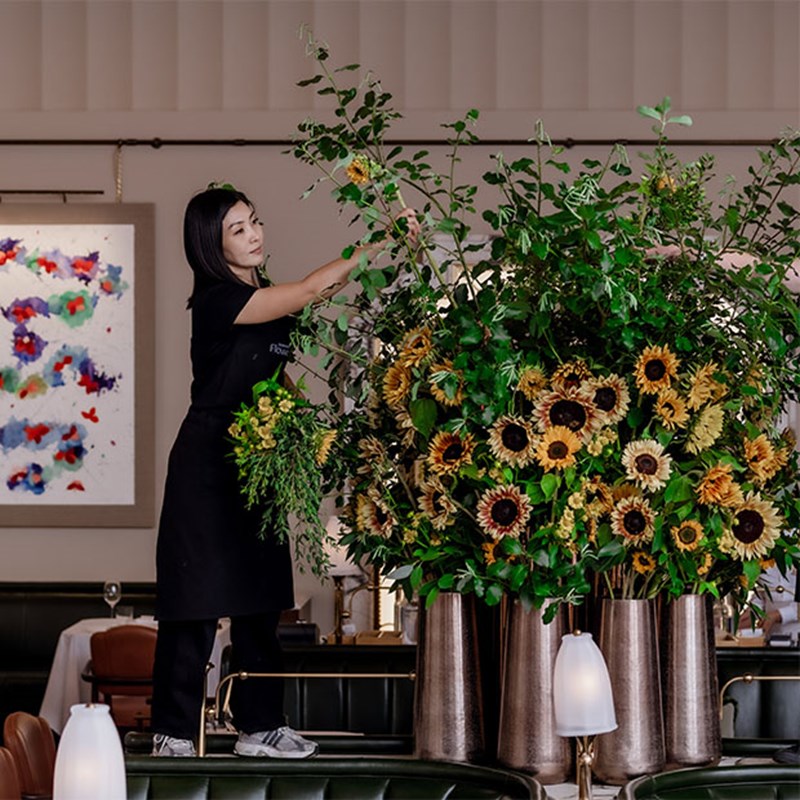 A florist in black uniform reaches up to refine a dramatic sunflower and greenery display, ensuring every stem is perfectly placed in the grand Claridge’s dining space.