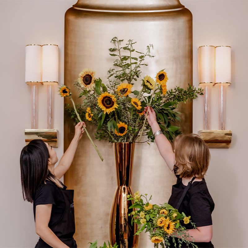 Two florists make final adjustments to a striking sunflower arrangement in a tall, golden vase, set against an elegant gold wall feature illuminated by warm sconces.