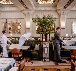 A grand dining space at Claridge’s, where staff in crisp uniforms gracefully prepare tables, flanked by plush green banquettes and a striking floral arrangement of sunflowers under stained-glass skylights.