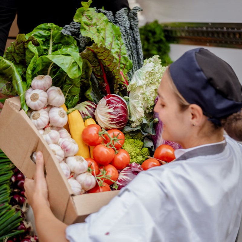A close-up of a chef carefully lifting a box filled with vibrant fresh vegetables, including tomatoes, garlic, rainbow chard, and Romanesco cauliflower, destined for the kitchen.