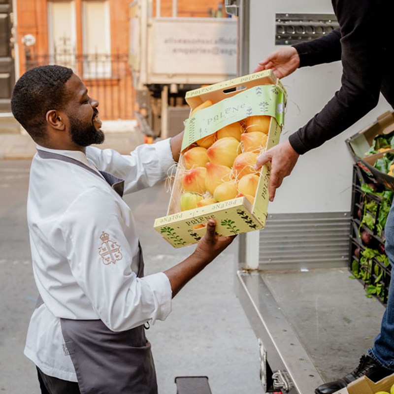 A delivery driver passes a wooden crate brimming with fresh apples to a Claridge’s chef, ensuring the finest ingredients make their way from market to menu.