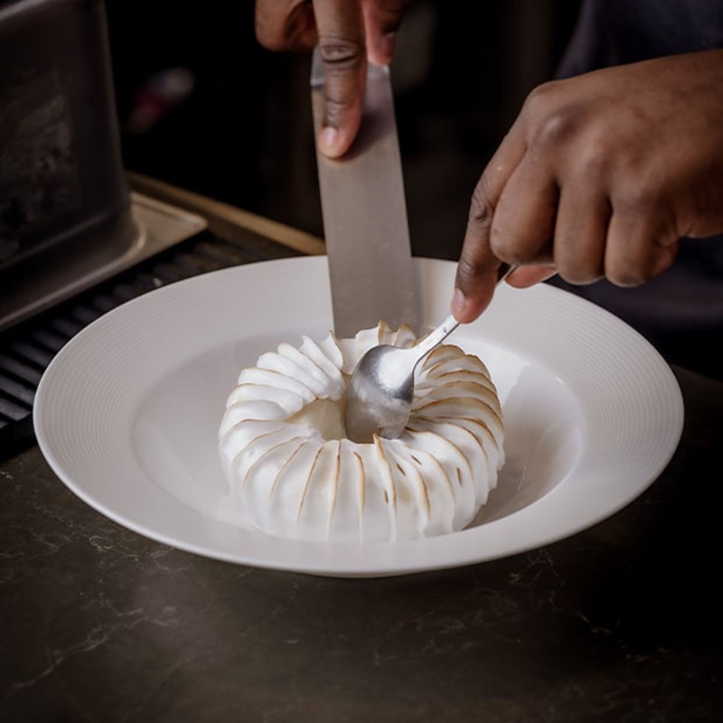 A close-up of a pastry chef’s hands as they expertly slice into a meticulously piped meringue ring, revealing its delicate layers on a pristine white plate.