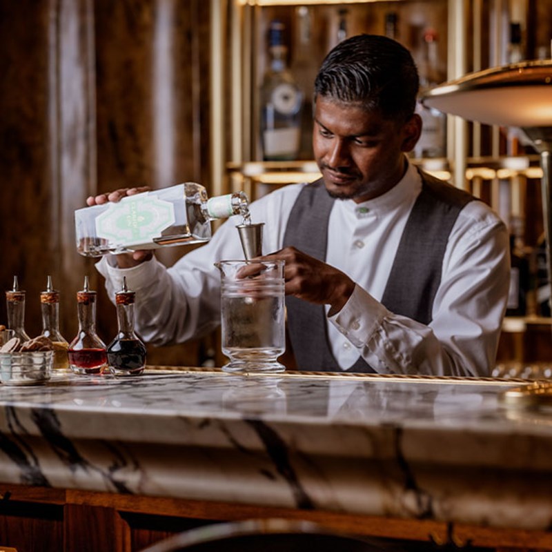 A bartender in a crisp white shirt and waistcoat carefully pours gin through a jigger into a mixing glass, surrounded by an elegant array of bitters and garnishes.