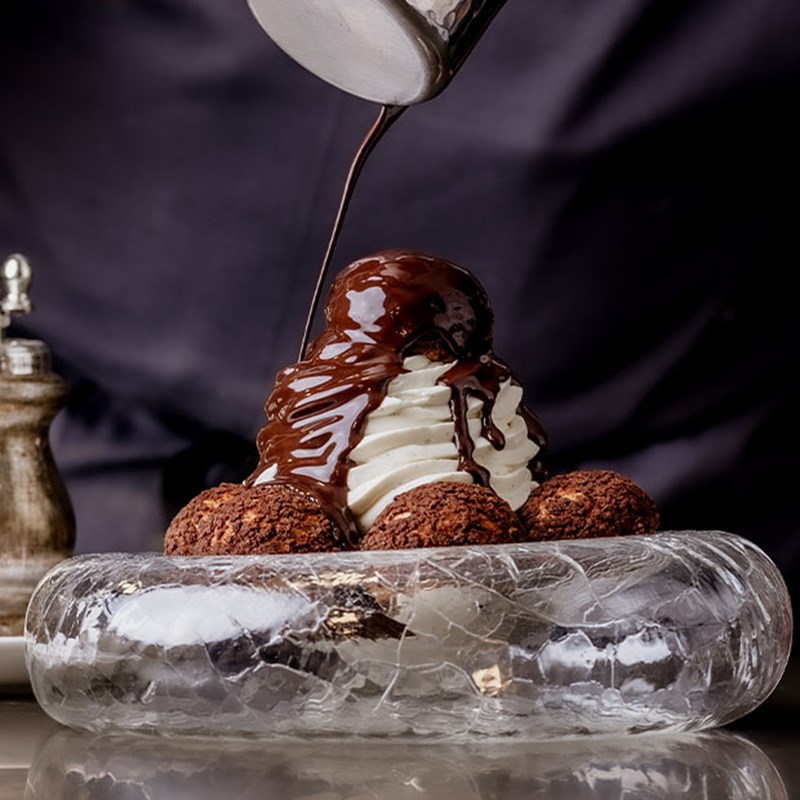 A sumptuous dessert of choux pastries filled with cream is crowned with a glossy cascade of rich, dark chocolate, served on an intricately textured glass plate.