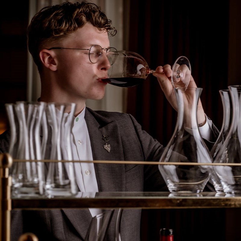 A young sommelier in refined attire savors a glass of deep-hued red wine, elegantly framed by a collection of delicate glassware on a golden trolley.