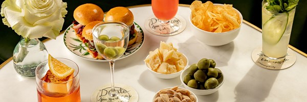 A marble table set with cocktails, sliders, olives, crisps, almonds, cheese chunks, and a white rose in a vase.